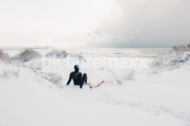It is cold and winter. A surfer sits on a snowy beach with a surfboard - watching the waves. A winter day with a surfer in a wetsuit.