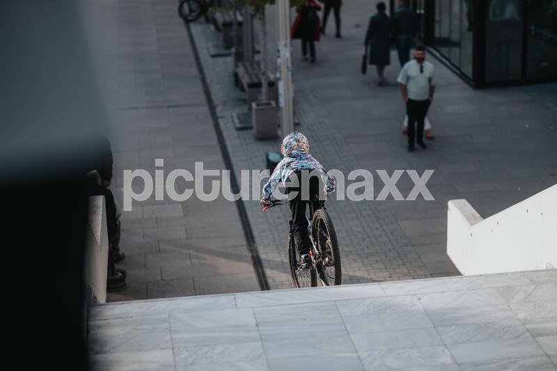 Ein kleines Kind fährt mit einem Fahrrad auf einem städtischen Gehweg und trägt einen gemusterten Helm.