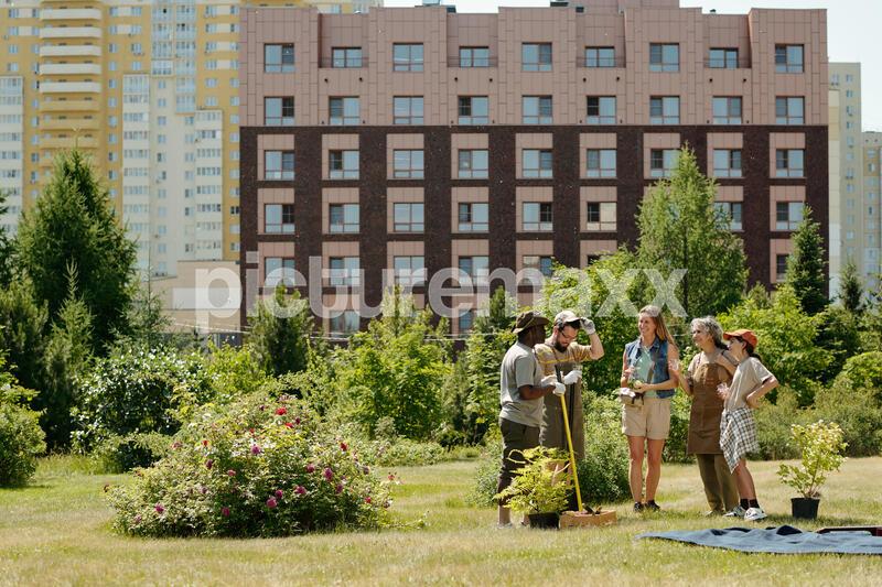 Group of diverse young adults and teenagers standing together in urban park, holding gardening tools and plants, engaging in community gardening activity with residential buildings behind