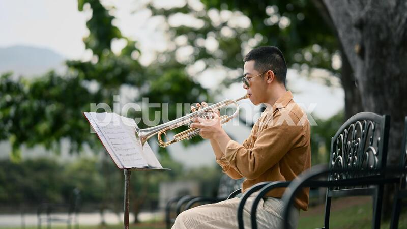 Young man playing trumpet outdoors while reading sheet music in a peaceful park.