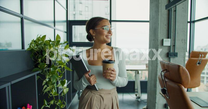 a confident young woman in an elegant outfit, holding a coffee cup and laptop, walking through a bright modern office space with large windows, stylish furniture, and green plants